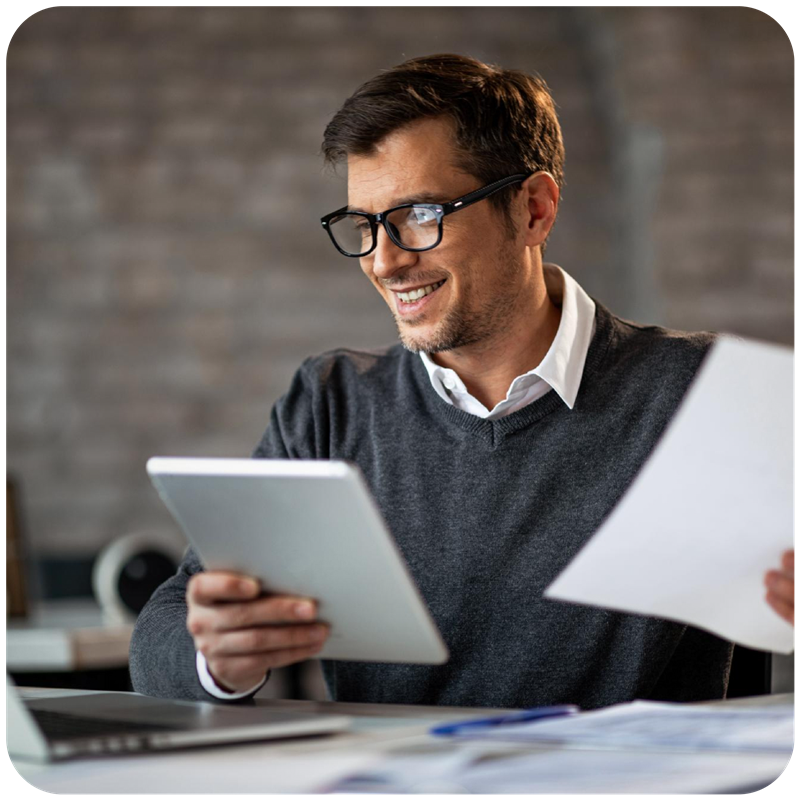 Smiling man wearing glasses, sitting at a desk while reviewing a document in one hand and holding a tablet in the other, representing a professional reviewing telecom expense management or telecom tools in a modern office setting.