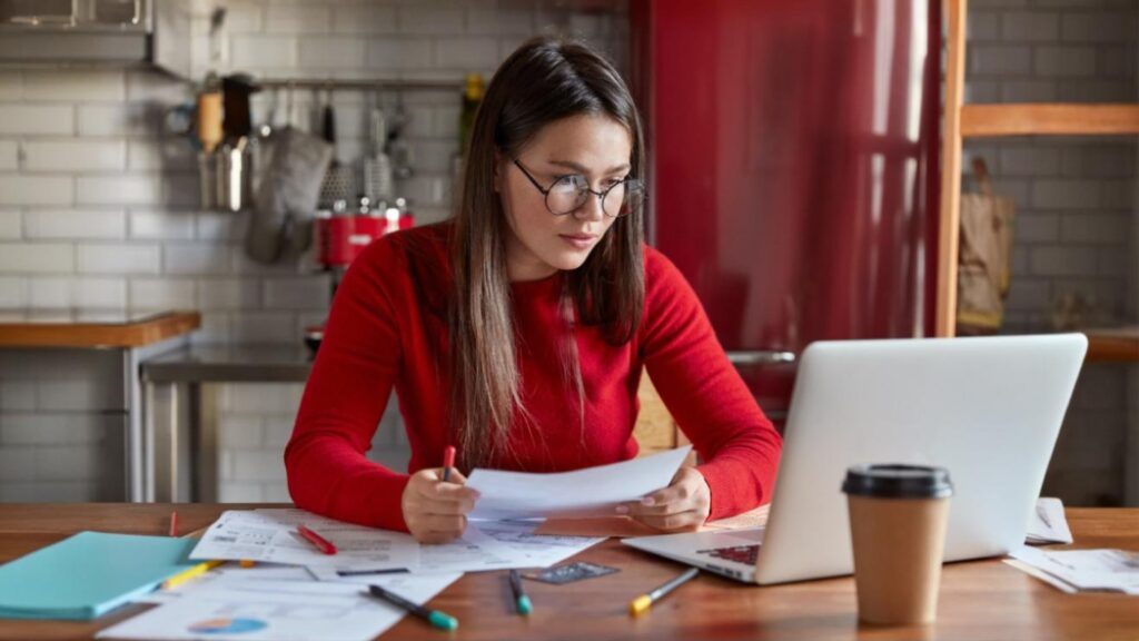 Woman in red sweater reviewing telecom billing documents at a laptop, with charts and a coffee cup on the table.