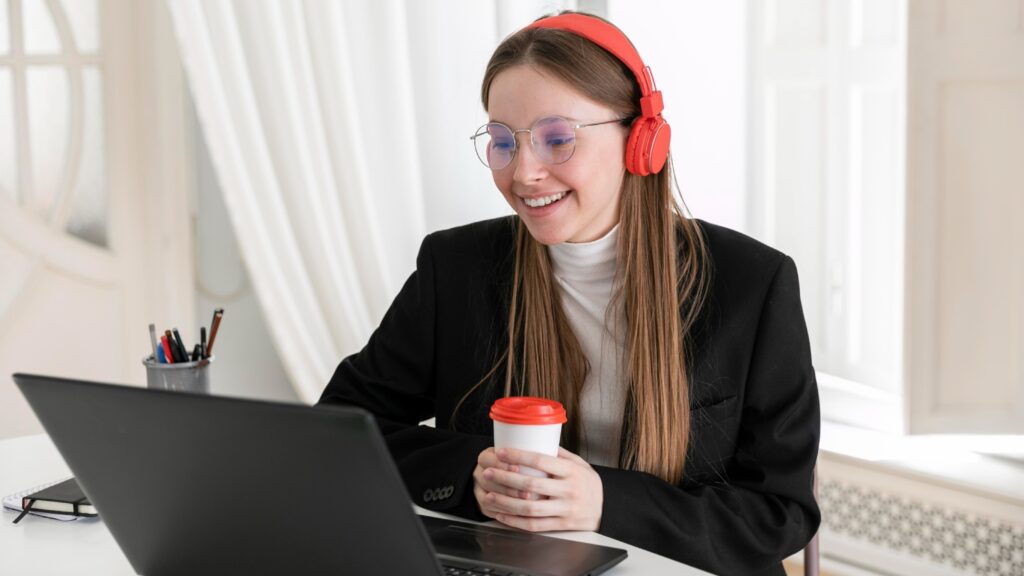 Young woman in a black blazer smiling during a video call, wearing red headphones and holding a coffee cup, with a laptop open in front of her—representing remote work using hosted phone systems.